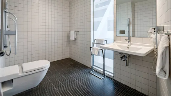A clean bathroom features a wall-mounted toilet, a large mirror above a sink, and white tiles. Towels hang on a rack, and the space is well-lit with modern fixtures.
