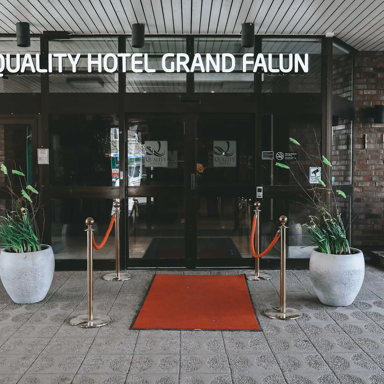 Glass entrance with red carpet leading to "QUALITY HOTEL GRAND FALUN." Flanked by potted plants; to the right, a colorful Dala horse statue stands against a brick wall.