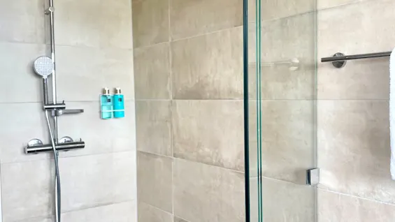 Modern shower area with a silver showerhead and glass door. Beige tiled walls, brown mosaic floor tiles; two blue bottles, white towel hanging on the right. No text visible.