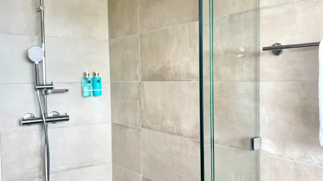 Modern shower area with a silver showerhead and glass door. Beige tiled walls, brown mosaic floor tiles; two blue bottles, white towel hanging on the right. No text visible.
