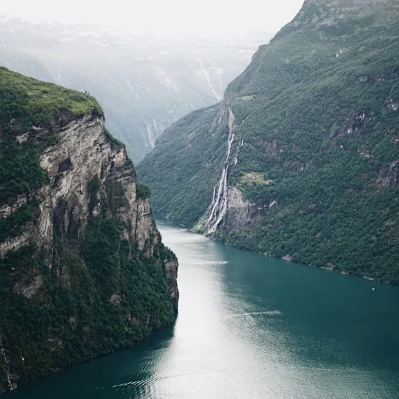 Naturskjønn utsikt over en dyp blå fjord som svinger seg mellom bratte, grønne fjell med en foss nedover fjellsiden.