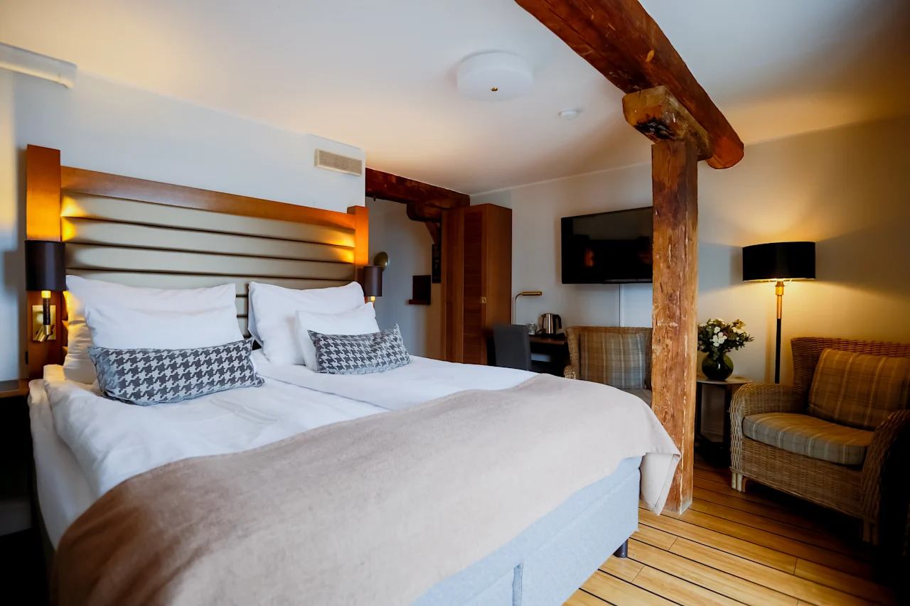 Et hyggeligt hotelværelse med en stor seng med houndstooth-puder, et vægmonteret TV og rustikke træbjælker.