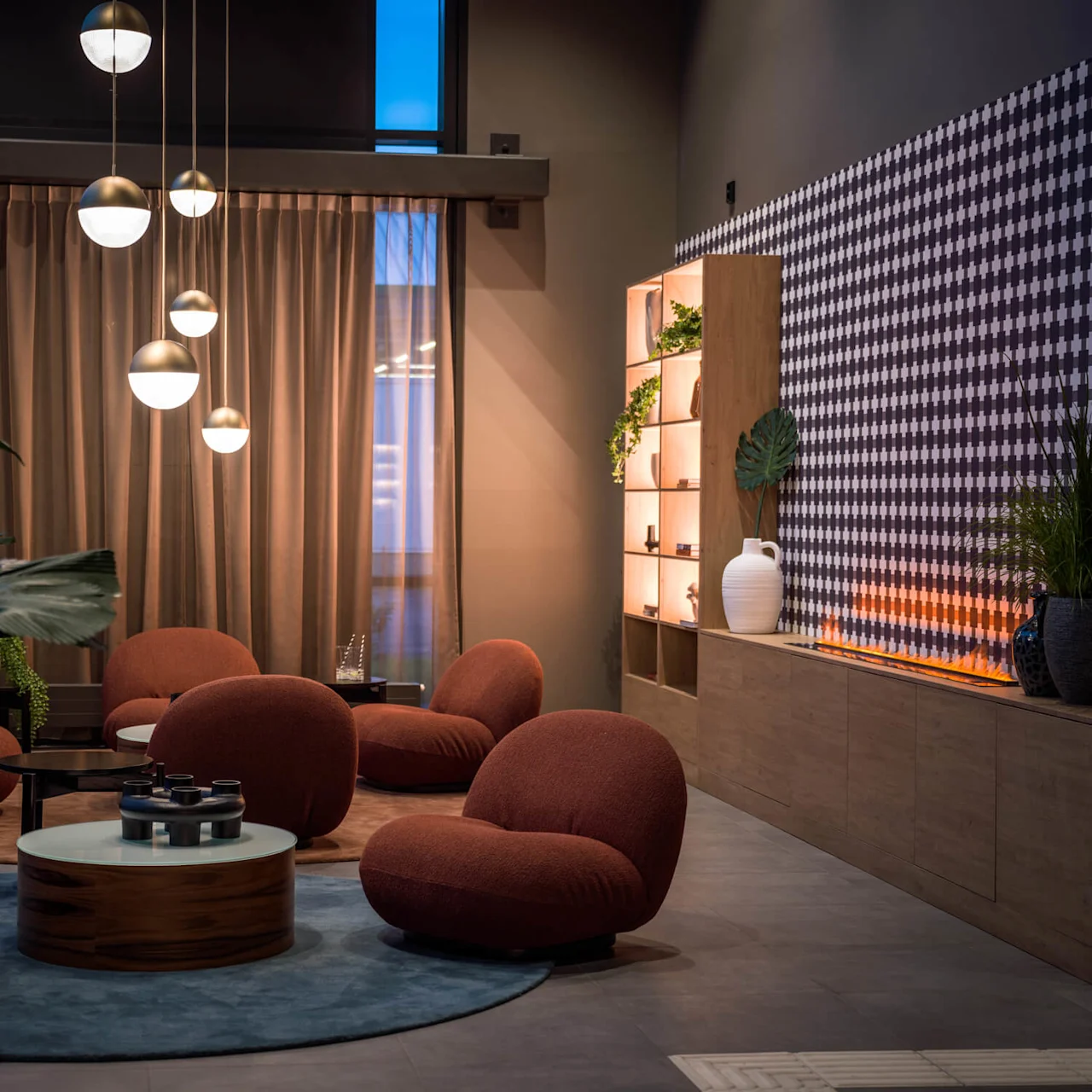 Runde, polstrede stoler står på et blått teppe i en moderne lounge. Hengende pendellamper lyser opp rommet, omgitt av en mønstret vegg, planter og opplyste hyller.