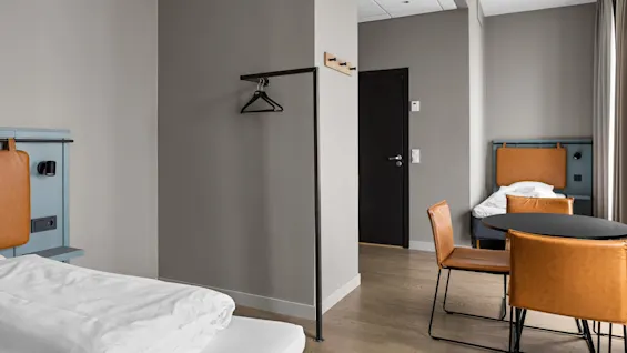 Minimalistinen hotellihuoneessa on nahkasomisteisia sänkyjä, pyöreä pöytä kahdella tuolilla ja metallivaatetanko, harmaiden seinien sekä puulattian ympäröimänä.