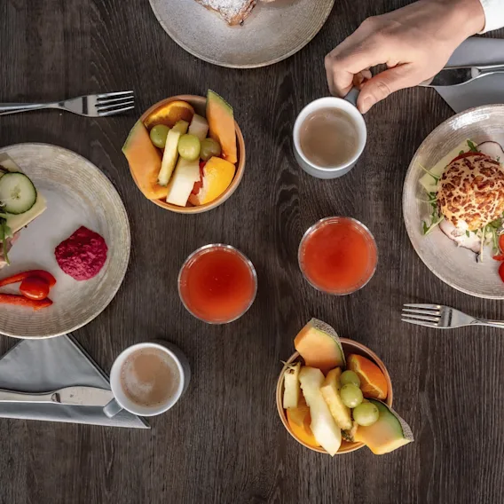 En bild ovanifrån av ett frukostbord i trä med två tallrikar med smörgåsar, två skålar med färsk frukt, två koppar kaffe och två glas juice.