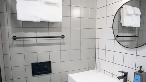 Hvite håndklær henger fra en svart stang på en grå flislagt vegg på et bad. Et rundt speil reflekterer håndklærne. En hvit vask har en dispenser merket "HAND WASH".