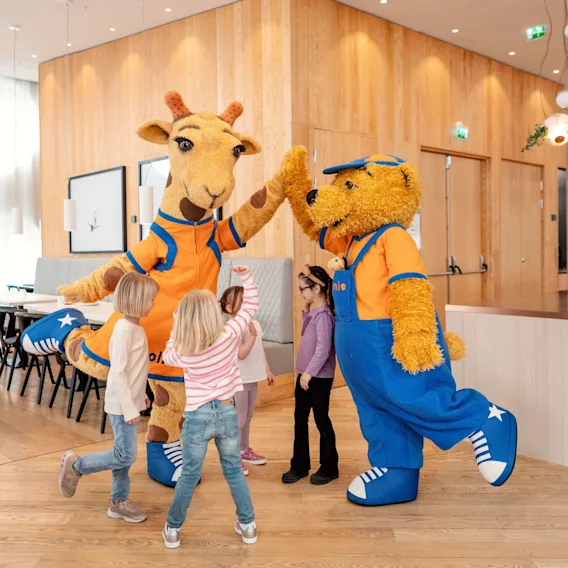 To maskoter, en sjiraff og en bjørn, gir high-five til barn i et trepanel-rom. Bjørnen har på seg klær med teksten "Lollo & Bernie". Lysende interiørbelysning styrker den livlige atmosfæren.