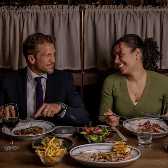En man i mörk kostym och en kvinna i en grön topp njuter av en middag vid ett restaurangbord fyllt med rätter som ryggbiff, pommes frites och skaldjur, medan de ler mot varandra.