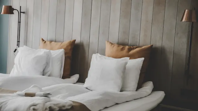 A neatly made double bed with white bedding and brown pillows rests against a wooden panel wall. Two wall-mounted lamps provide soft lighting, creating a cozy atmosphere.