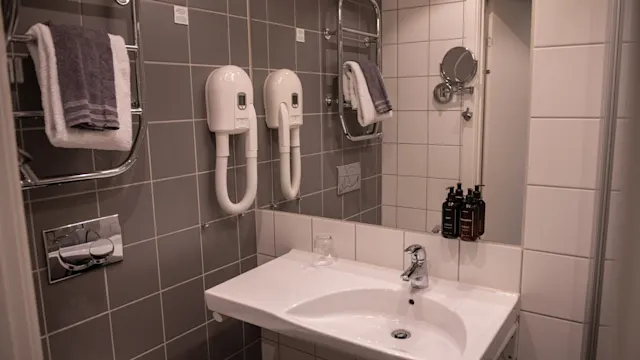 Ett badrum med två väggmonterade hårtorkar ovanför ett handfat. Kaklade väggar omger en handdukshållare, spegel och toalettartiklar. Handdukar hänger prydligt på hållare.