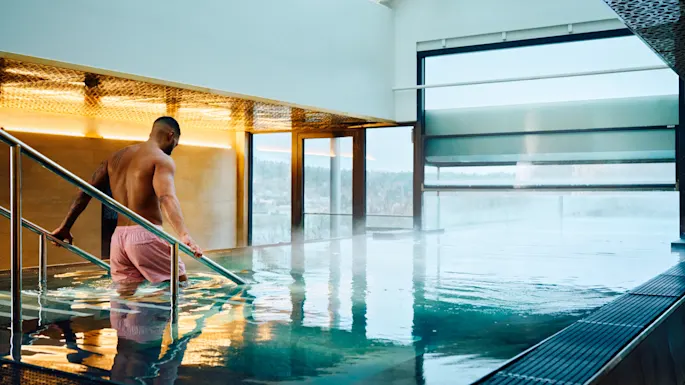 A person in pink shorts descends metal steps into an indoor pool, surrounded by large windows and modern architecture.
