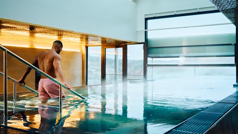 A person in pink shorts descends metal steps into an indoor pool, surrounded by large windows and modern architecture.