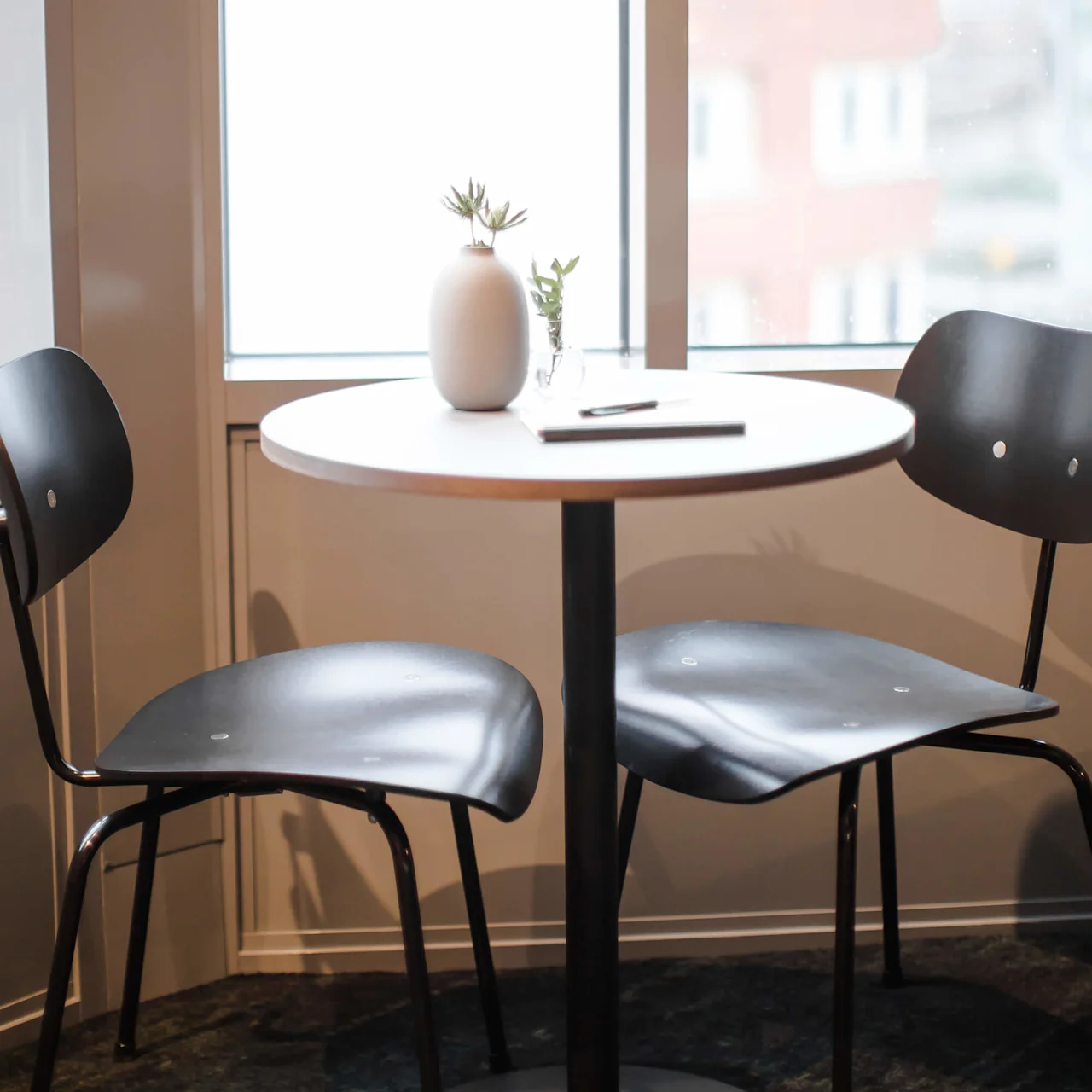 A small round table with a white vase and plant sits between two black chairs, in a room with a large window showing a blurred cityscape outside.