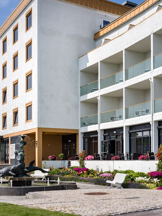 The exterior of Quality Hotel Grand Larvik, featuring a white building with balconies, a vibrant flower garden, and a fountain.