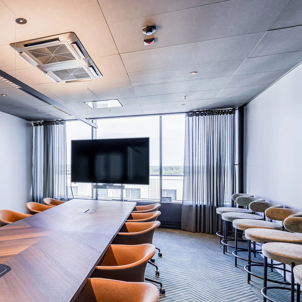 Conference table is surrounded by orange chairs, situated in a modern meeting room with large windows, a television, and several bar stools against one wall. No text is present.  