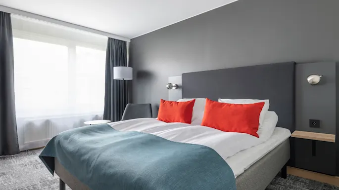 En seng med røde puter og blå sengeteppe er i et moderne rom. Det har grå vegger og et vindu med mørke gardiner og en lampe.