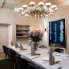 A dining table is elegantly set with folded napkins and wine glasses, surrounded by black chairs. A chandelier hangs overhead in a modern room with artwork on the wall.