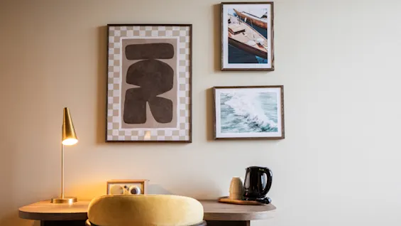 Skrivbord med en gul stol, en lampa, en vattenkokare och två inramade foton på en beige vägg. En stor abstrakt konst hänger i närheten.