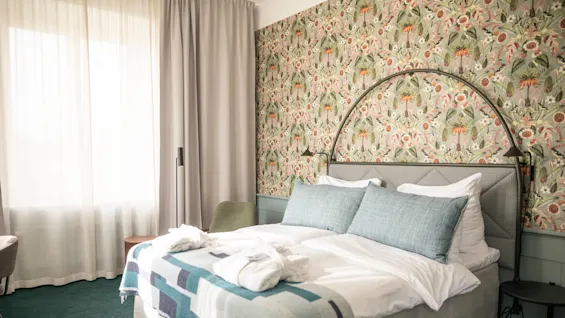 Säng klädd med vita lakan och blå kuddar, mot blommig tapet. Solljus filtrerar genom gardiner och lyser upp ett mysigt hotellrum med en mjuk grön matta.