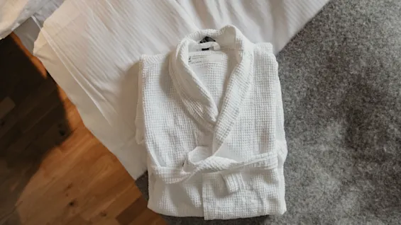A folded white bathrobe rests on a bed, with a gray blanket nearby and wooden flooring partially visible.