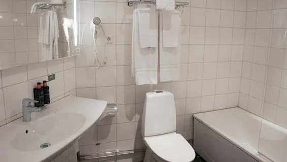 A clean, tiled bathroom with a toilet, sink, and bath, reflected in a mirror with hanging towels and a wall-mounted hairdryer.