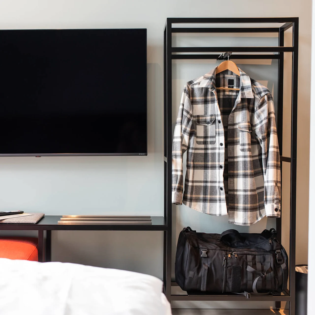 A plaid shirt hangs on a rack beside a black duffel bag; a flat-screen TV is on the wall in a minimalistic room.