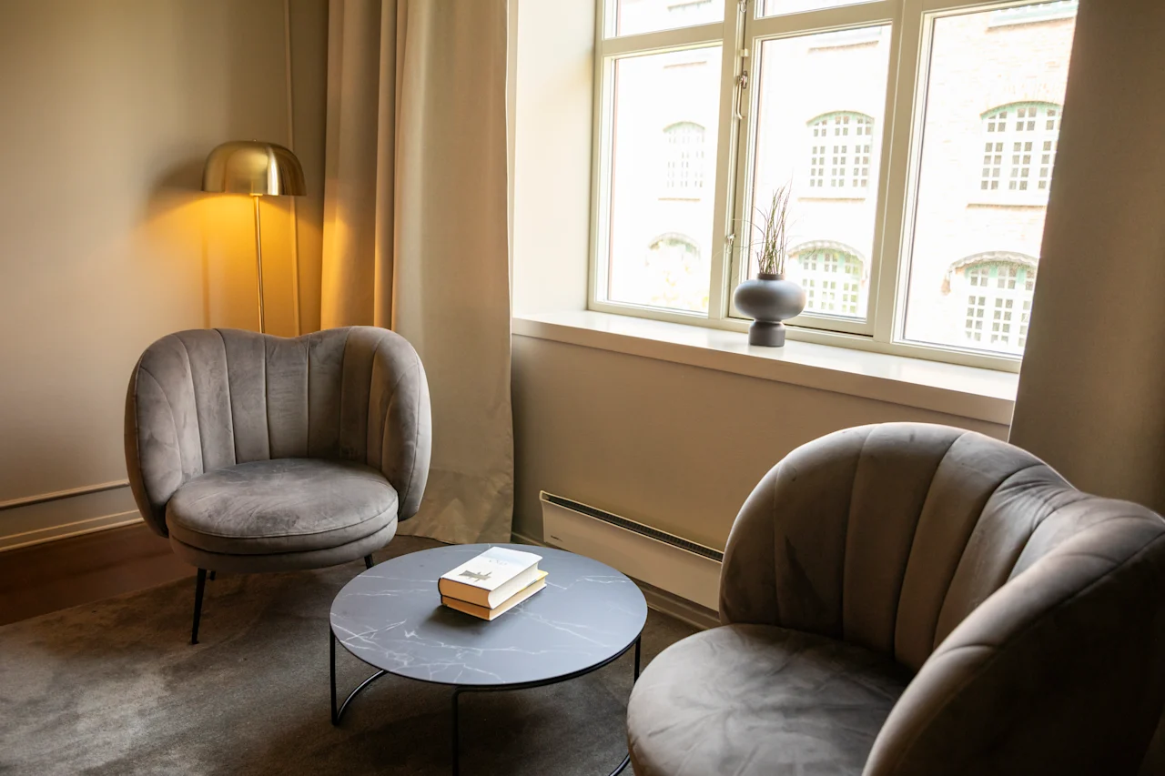 To grå lenestoler vender mot et rundt svart kaffebord som holder en bok, i et mykt opplyst rom med et vindu, beige gardiner, en gyllen lampe og potteplante.