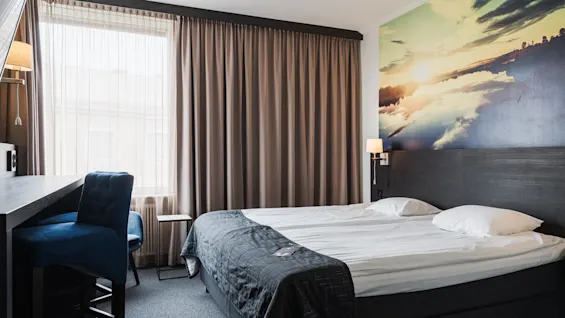 En snyggt bäddad säng med kuddar, i ett hotellrum med en stor väggmålning av en solnedgång över en sjö. En blå stol och ett skrivbord står vid fönstret.
