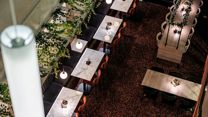 Marble-topped tables line a contemporary dining area, paired with orange chairs, surrounded by hanging greenery and warm globe lights, creating an inviting restaurant ambiance.