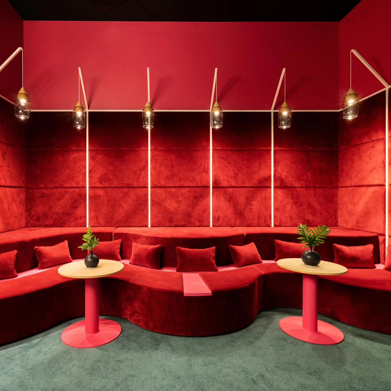 Curved red velvet seating area adorned with matching cushions surrounded by red padded walls and pendant lights; small round tables with plants accent the space, set against a green carpet.