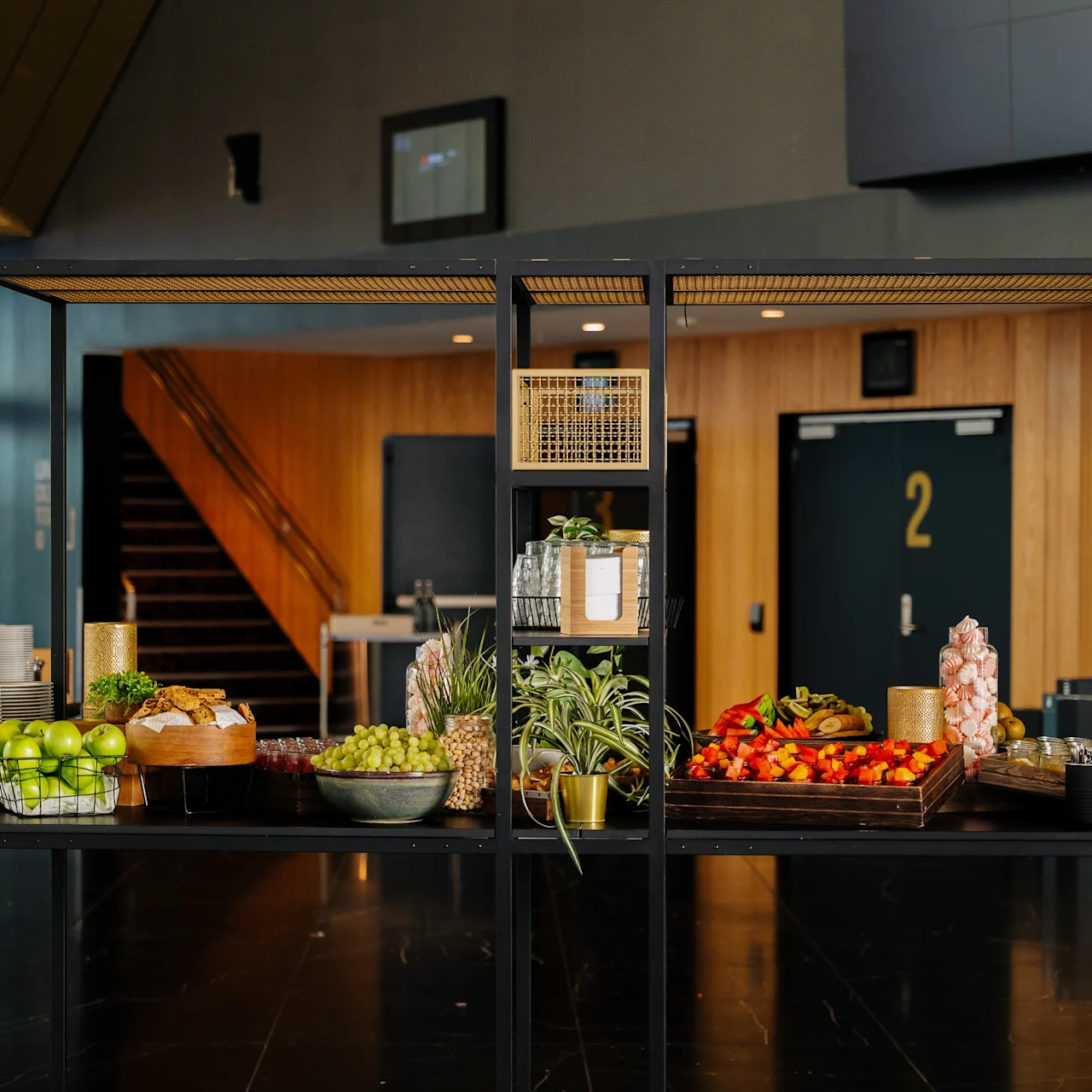 A black metal buffet table displays fruits, plants, and beverages in a modern indoor setting with wood-paneled walls, a staircase, and visible door labeled "2."