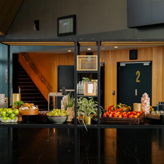 Et buffetbord av svart metall viser frukt, planter og drikkevarer i et moderne innemiljø med panelvegger, en trapp og en synlig dør merket "2."