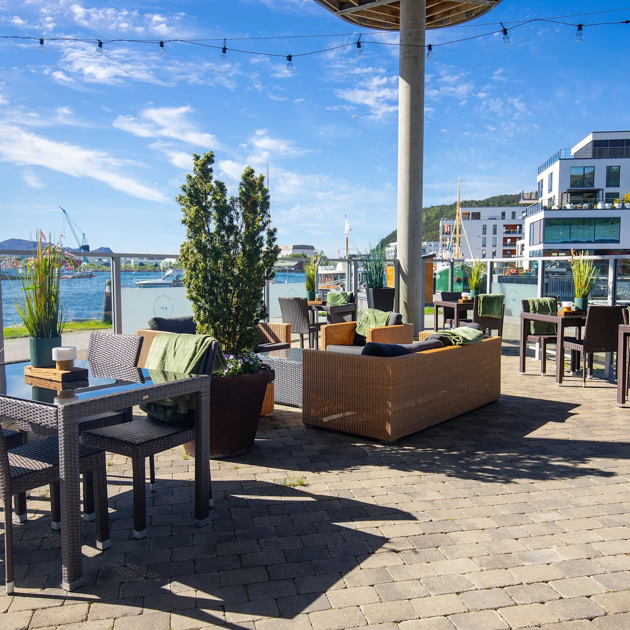 Utendørs kafépatio med rottingbord og stoler arrangert for servering, pyntet med planter og fargerike puter, med utsikt over en marina under en klar blå himmel. Synlige lysslynger gir stemning.