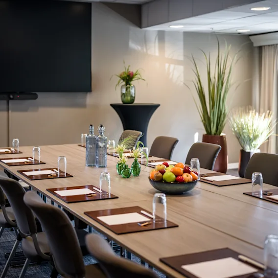 A modern conference room with a long table set for a meeting, featuring a large screen, fruit, and plants.
