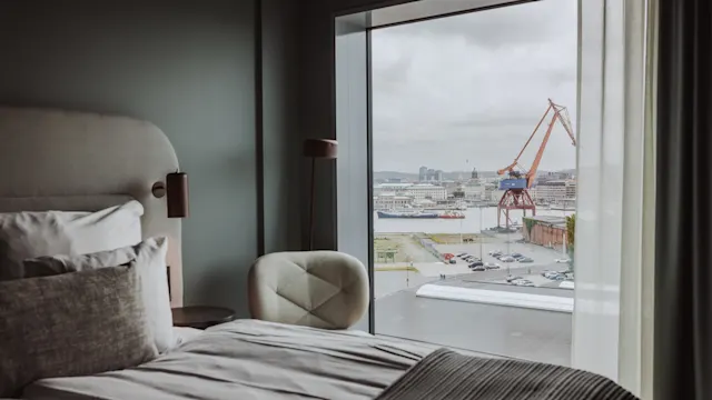 En seng med gråt og hvidt sengetøj er placeret ved et stort vindue med udsigt over en havn med en rød kran. Værelset har moderne indretning.