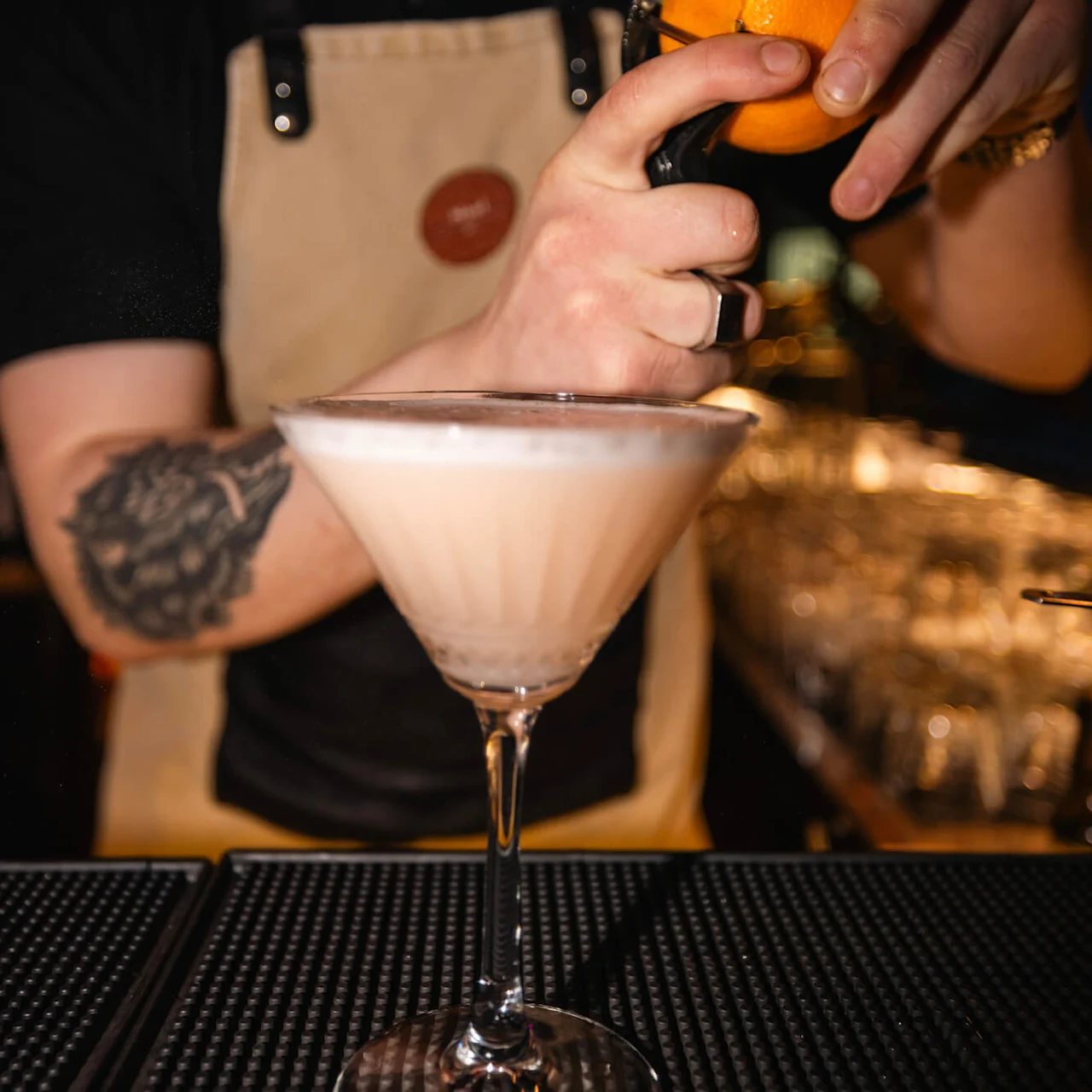 Bartender skalar en apelsin över ett skummande cocktailglas. Han bär ett förkläde och står i en svagt upplyst bar med glas i bakgrunden.