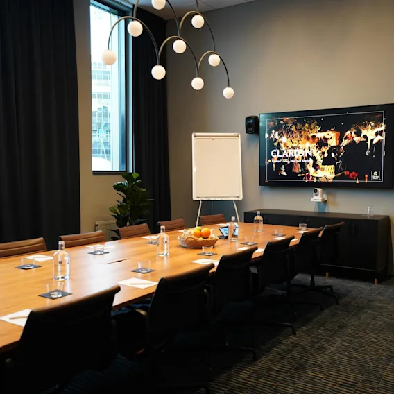 A long conference table is set with water bottles and notepads beneath a bright overhead light. A screen displaying "Clarion" features festive imagery in a modern meeting room.
