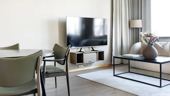 A flat-screen TV is mounted on a wall unit in a modern living room. Nearby, a round dining table with chairs, a beige sofa, and a black coffee table with a vase of flowers complete the setting.