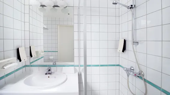 A clean, white-tiled bathroom with a sink, mirror, hand dryer, paper dispenser, and a shower corner with a showerhead and controls.