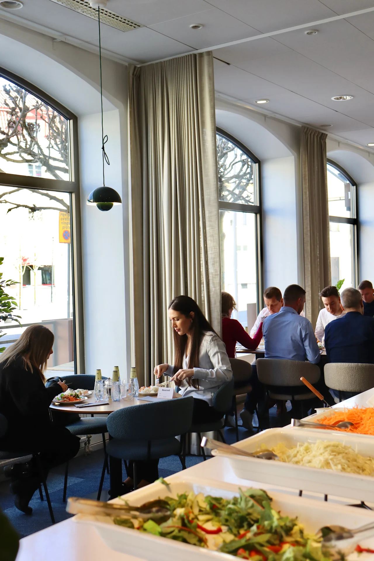 A dining area with people eating at tables near large windows; a salad bar in the foreground offers various ingredients. Sunlight illuminates the modern, bright space.
