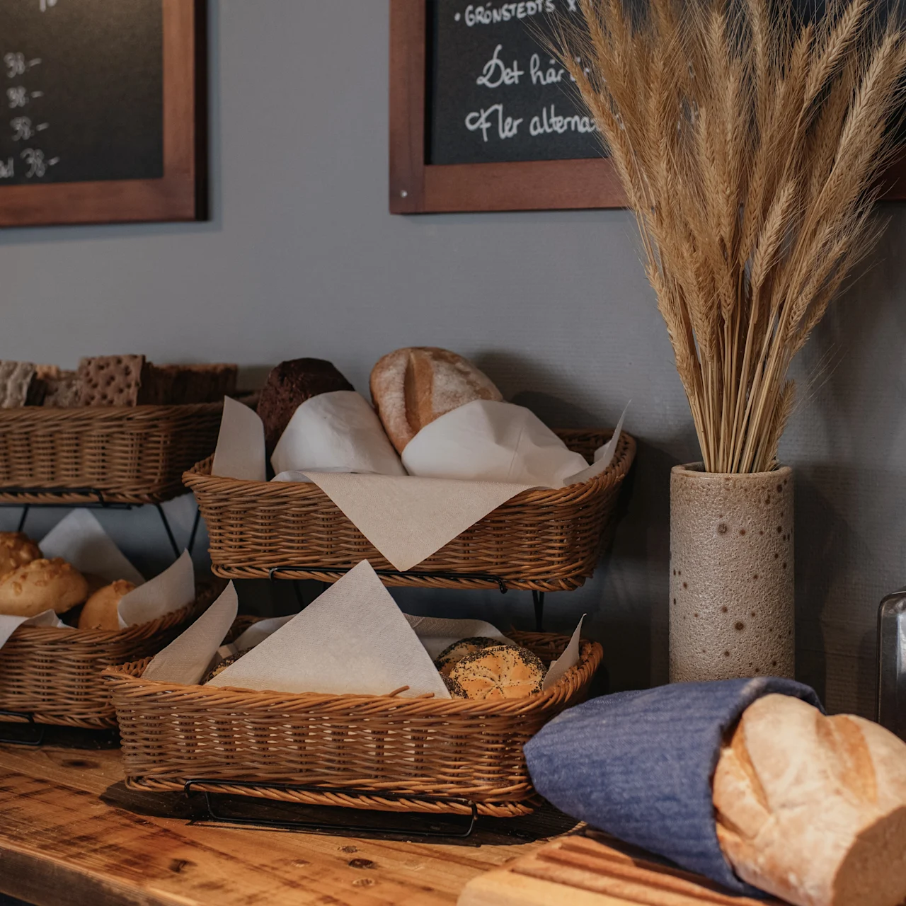Baskets of assorted bread rest on a wooden counter; a vase with dried wheat decorates the space. Chalkboards with menus hang on the wall.