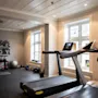 Løbebånd står ubrugt i et lyst fitnessrum med lyse vægge. Bag det hviler kettlebells og en træningsbold på gulvet tæt på en motiverende vægmaleri: "EVERY PERSON YOU KNOW USED TO BE A STRANGER. THE PERSON YOU ARE IS UP TO YOU."