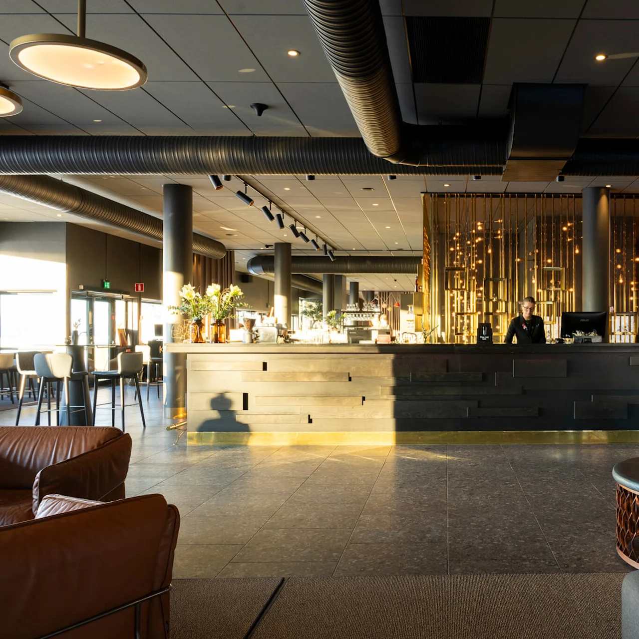 En person står bakom en stilren, upplyst hotellreception med varm belysning. I den rymliga lobbyn syns lädersoffor och dekorativa kuddar.