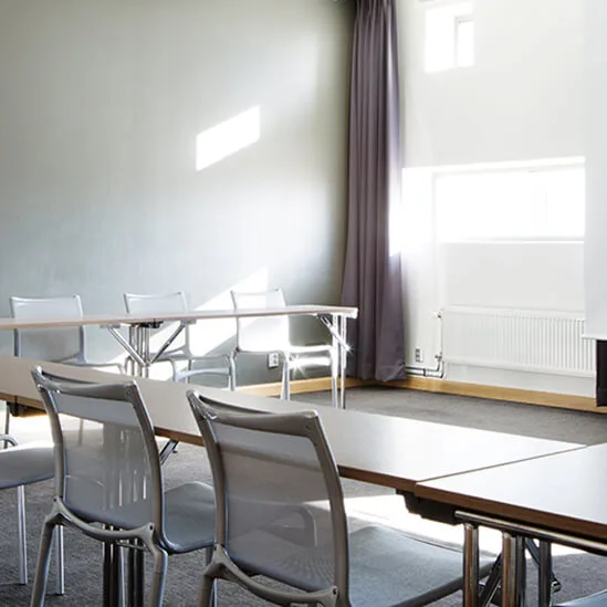 Meeting room with empty chairs arranged around rectangular tables. A large blank projection screen dominates the room, surrounded by light gray walls and large windows illuminating the space.