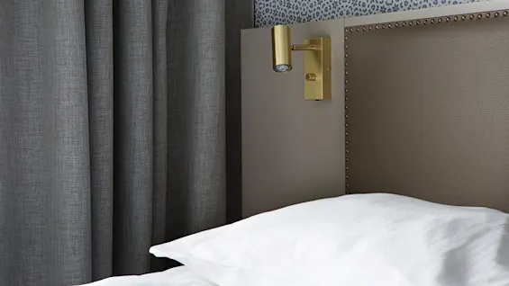 A neatly made bed with a white pillow, beside a studded headboard and a modern wall-mounted lamp, in a room with patterned wallpaper and grey curtains.