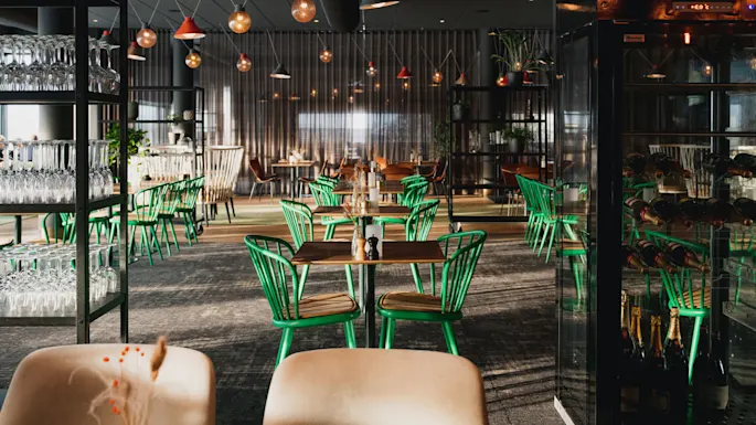 Bord med grønne stoler er plassert, omgitt av hyller med glass. Hengelamper henger over i en svakt opplyst moderne restaurant. En vinutstilling er på høyre side.