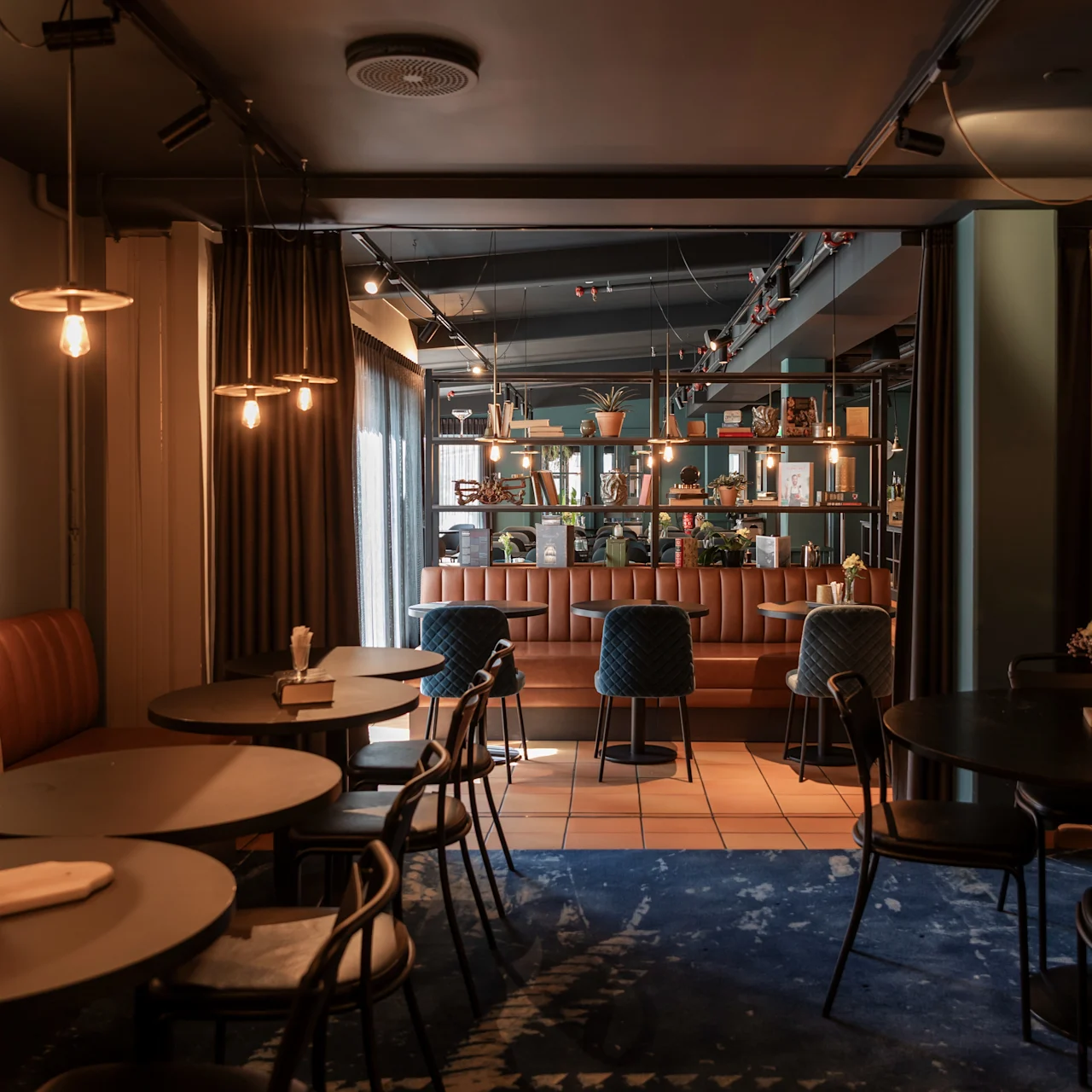 Hyggelig café med runde borde og stole på et mønstret blåt tæppe. En bardisk smykket med planter og lys strækker sig i baggrunden. Dæmpet, varm belysning skaber en indbydende atmosfære.