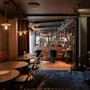 Hyggelig café med runde borde og stole på et mønstret blåt tæppe. En bardisk smykket med planter og lys strækker sig i baggrunden. Dæmpet, varm belysning skaber en indbydende atmosfære.
