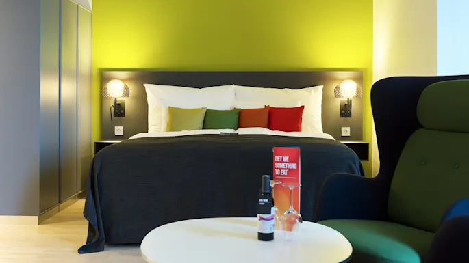 A bed with colorful pillows stands against a lime-green wall in a modern room. A table holds wine and a card saying, "GET ME SOMETHING TO EAT."