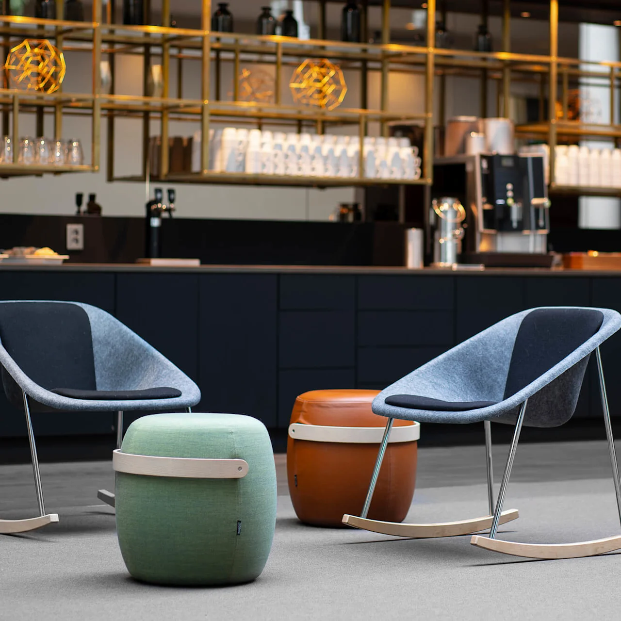 Rocking chairs rest beside cylindrical poufs in modern lounge; shelves with glasses and geometric lights, counter with coffee machine create a cozy cafe setting.